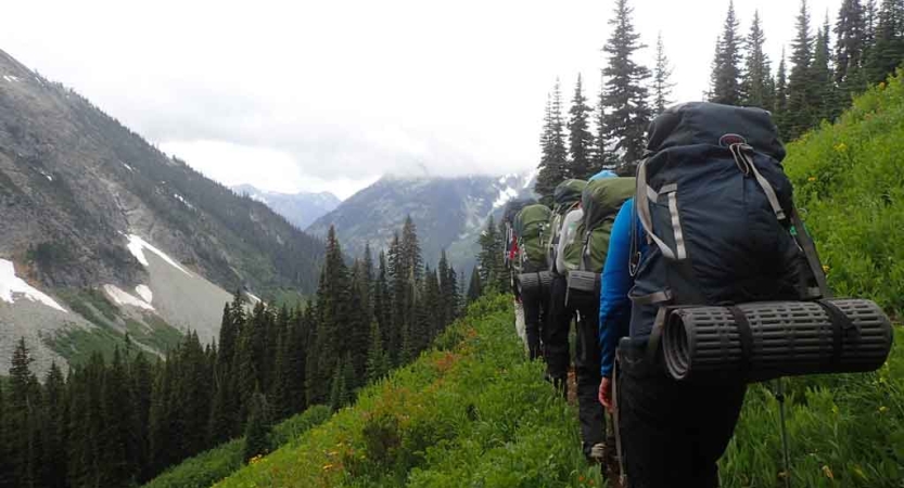 a group of students wearing backpacks hike along a grassy trail with mountains in the distance